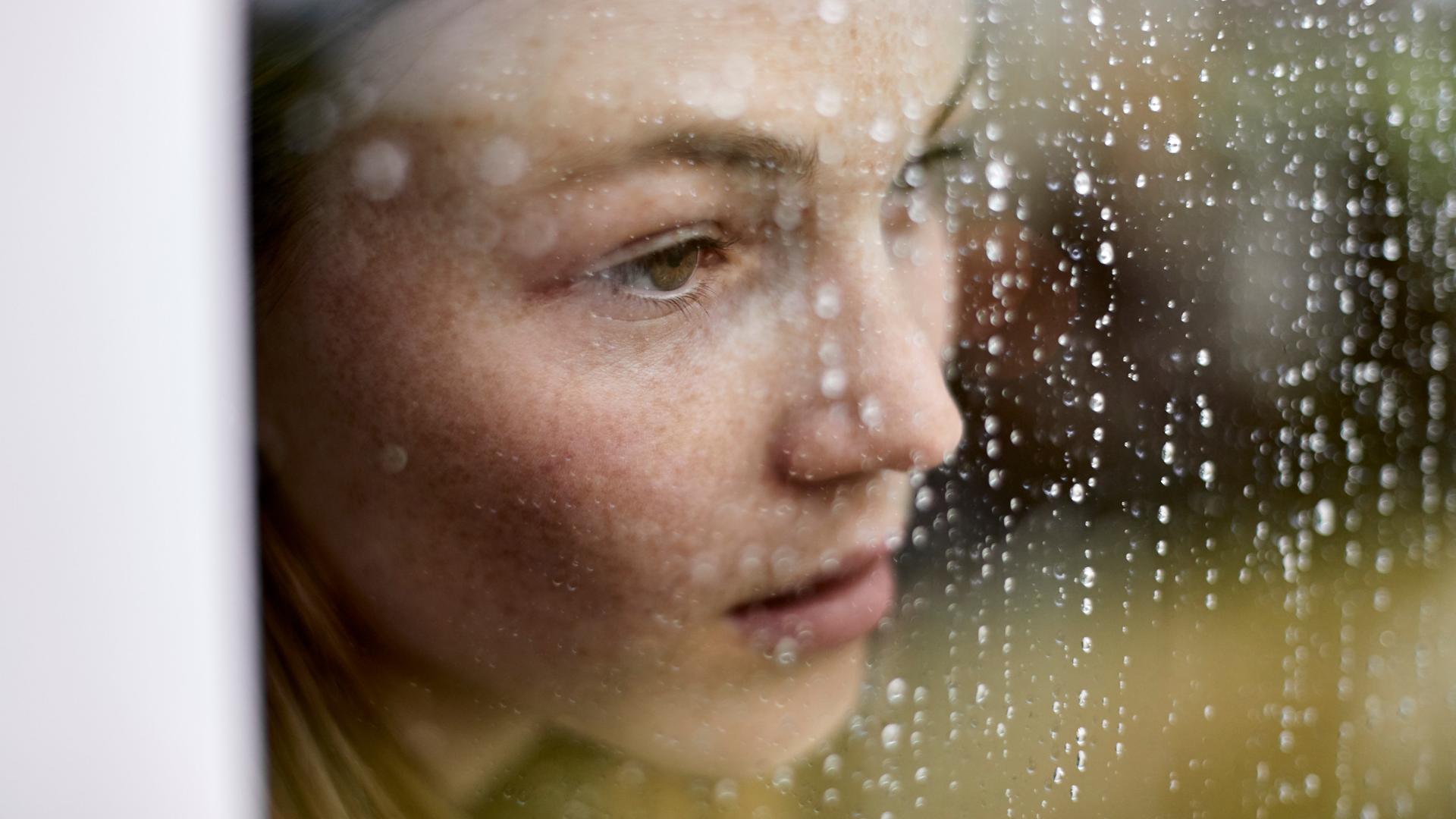 Eine junge Frau schaut traurig durch ein Fenster auf dem Regen sichtbar ist.