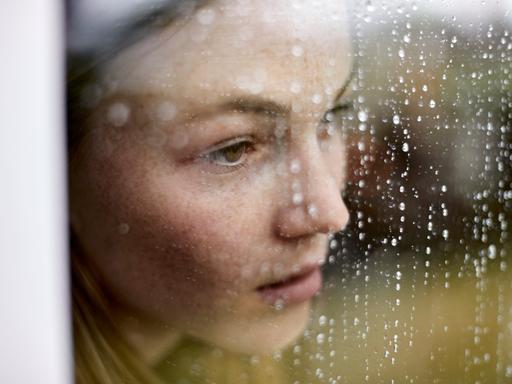 Eine junge Frau schaut traurig durch ein Fenster auf dem Regen sichtbar ist.