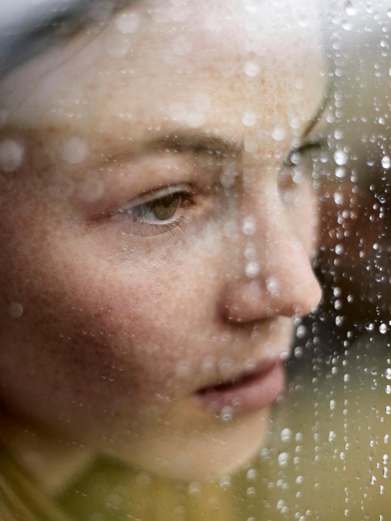 Eine junge Frau schaut traurig durch ein Fenster auf dem Regen sichtbar ist.