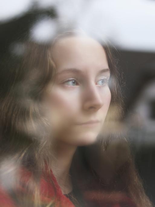 Eine junge Frau steht am Fenster, sie blickt raus, in der Scheibe spiegelt sich ein Haus.
