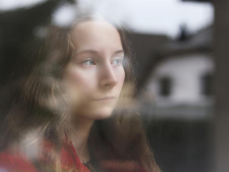 Eine junge Frau steht am Fenster, sie blickt raus, in der Scheibe spiegelt sich ein Haus.