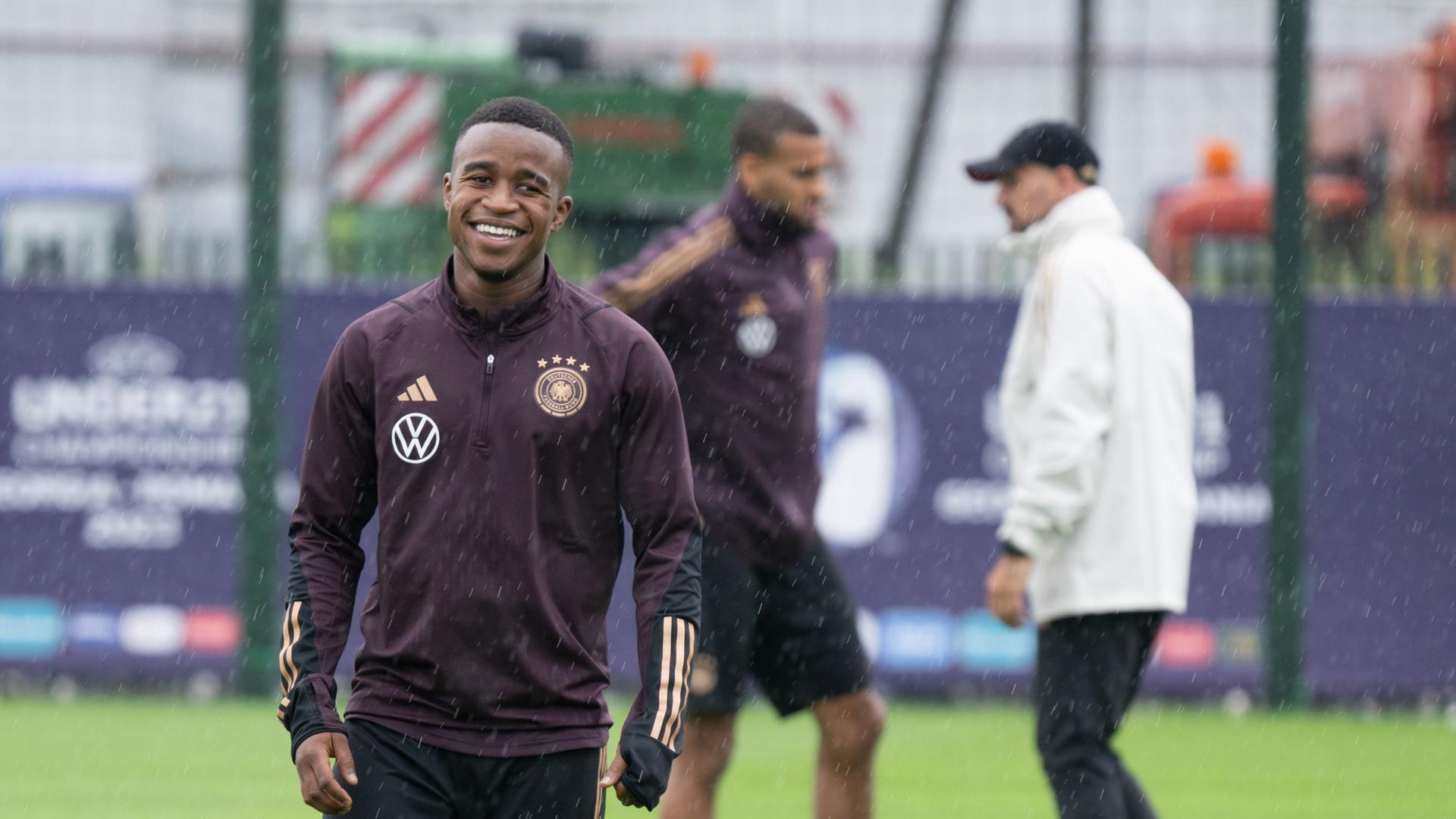 Youssoufa Moukoko steht im Trainingsanzug auf dem Spielfeld und lächelt in die Kamera.