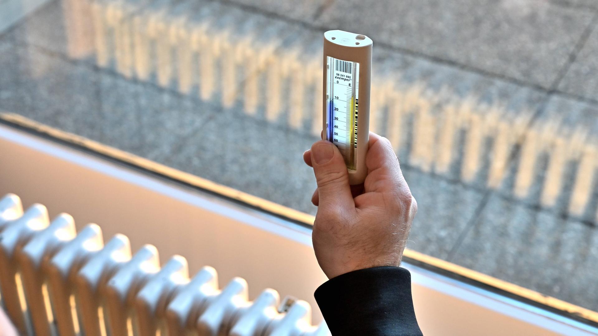 Vor einem Fenster hängt ein Heizkörper, ein Mann hält ein Fernablesegerät gegen das Licht sodass man die Thermometer-Anzeige lesen kann.