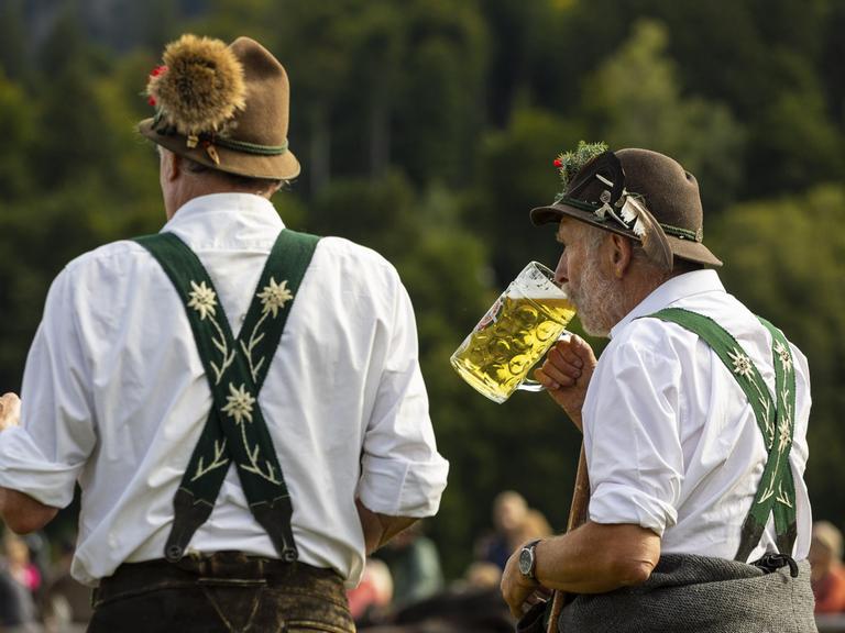 Zwei Männer in traditioneller Kleidung stehen beim Almabtrieb. Einer trinkt Bier aus einem großen Glas.