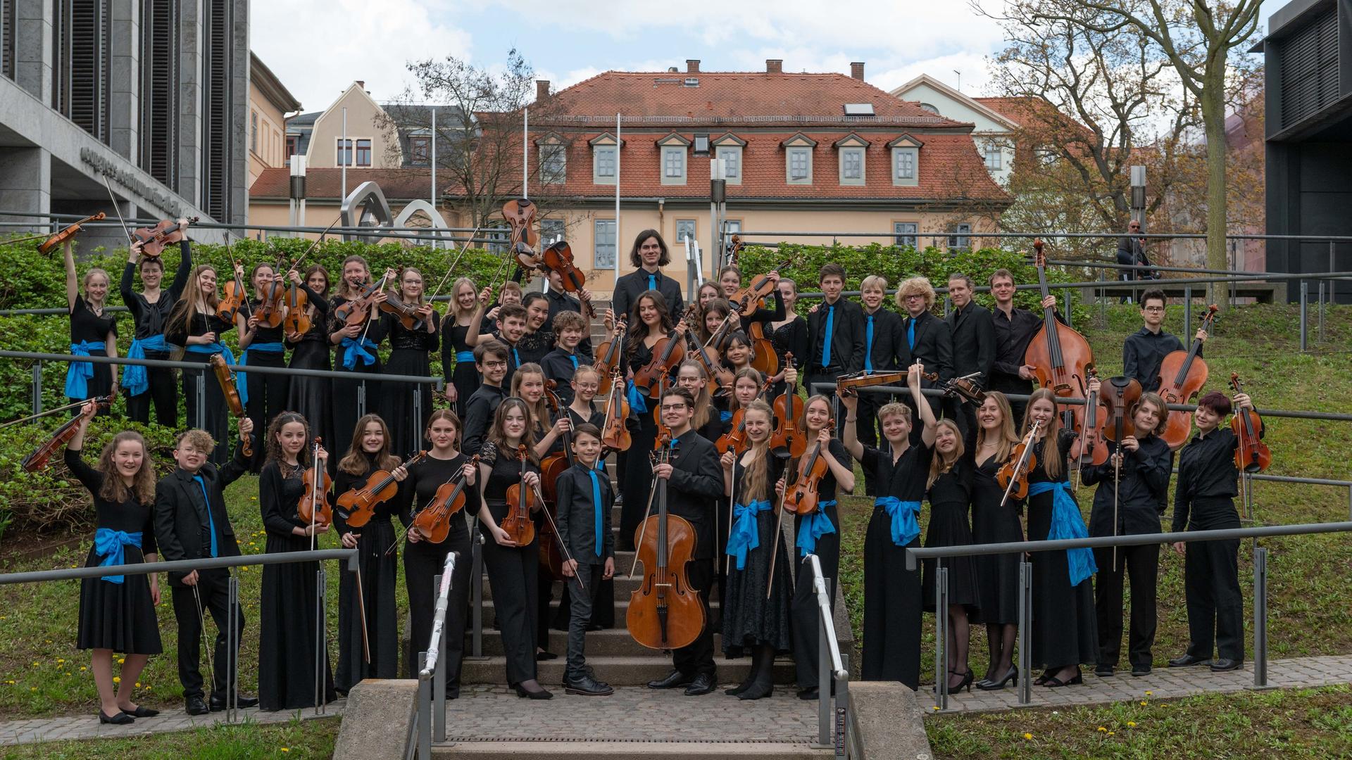 Die jungen Musikerinnen und Musiker der Deutschen Streicherphilharmonie stehen in Konzertkleidung mit ihren Instrumenten auf einer Freilufttreppe.