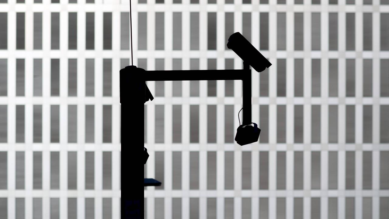 Die dunklen Umrisse einer Überwachungskamera – im Hintergrund ein graues Gebäude mit zahlreichen Fenstern
