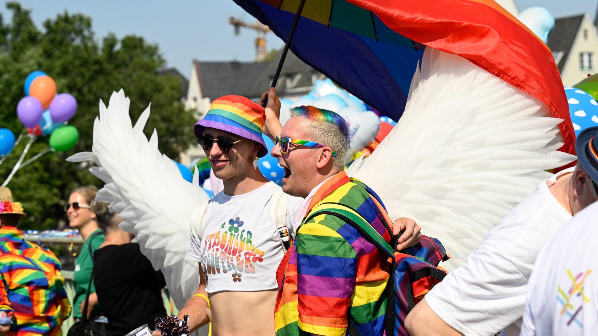 Zwei verkleidete Männer bei der Pride-Parade in Köln. Einer von den Männern hat Kleidung in Regenbogen-Farben an. Einer trägt weiße Flügel wie ein Engel.