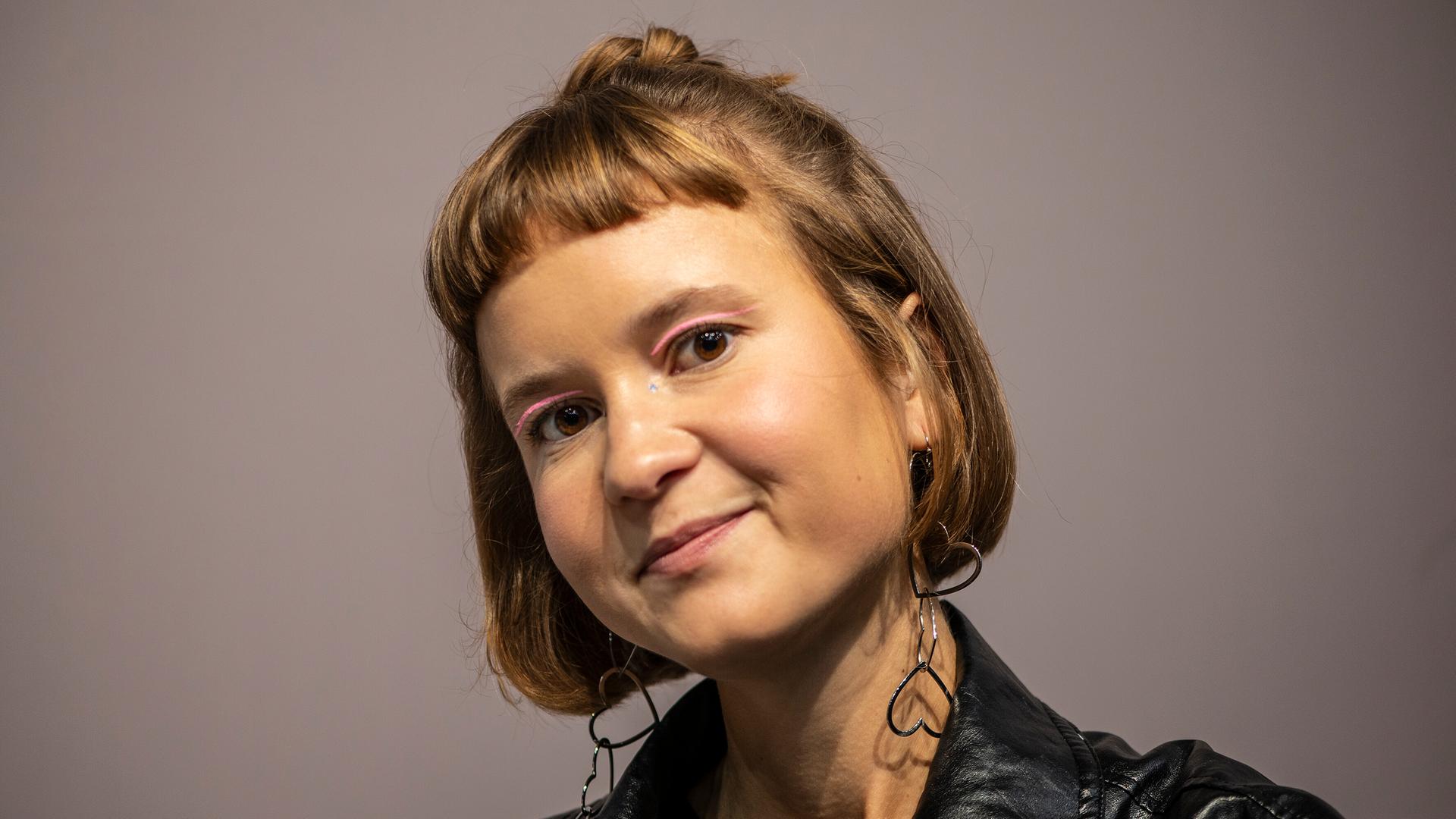 Caroline Wahl trägt eine schwarze Lederjacke, rosa Lidschatten und Ohrringe in Herzform. Sie schaut freundlich in die Kamera.