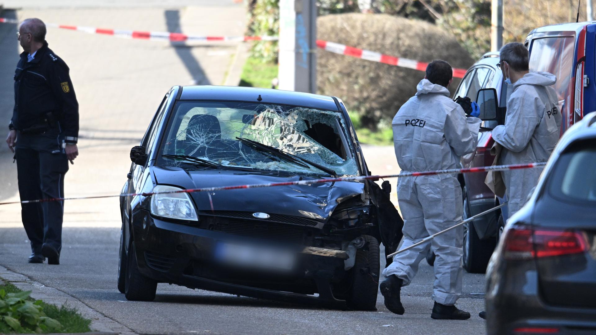 Der schwer beschädigte schwarze Kleinwagen steht auf einer Straße. Davor Flatterband und Ermittler in weißen Schutzanzügen. 