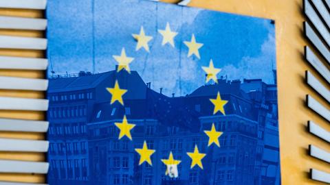 Brüssel: Drei Fahnen der Europäischen Union und gegenüberliegende Gebäude spiegeln sich im EU-Logo an der Außenfassade des Berlaymont-Gebäudes.