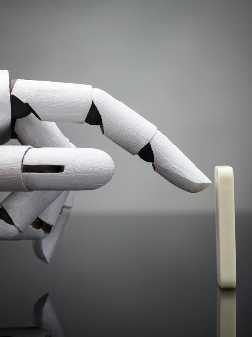 Eine Roboterhand ist kurz davor, einen aufgestellten Dominostein anzutippen. Eine Roboterhand ist kurz davor, einen aufgestellten Dominostein anzutippen.