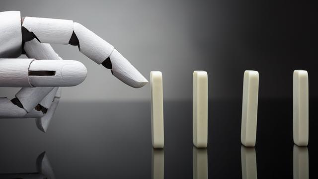 Eine Roboterhand ist kurz davor, einen aufgestellten Dominostein anzutippen. Eine Roboterhand ist kurz davor, einen aufgestellten Dominostein anzutippen.