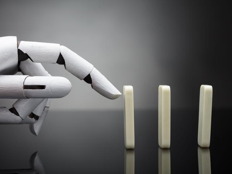 Eine Roboterhand ist kurz davor, einen aufgestellten Dominostein anzutippen. Eine Roboterhand ist kurz davor, einen aufgestellten Dominostein anzutippen.