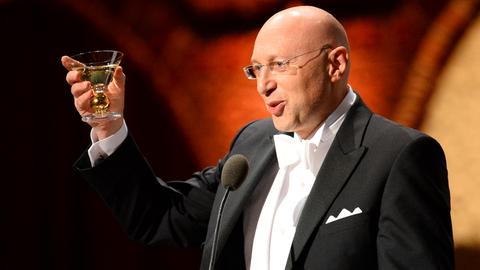 Stefan Hell steht im Smoking hinter einem Mikrofon und hebt sein Sektglas in die Höhe.