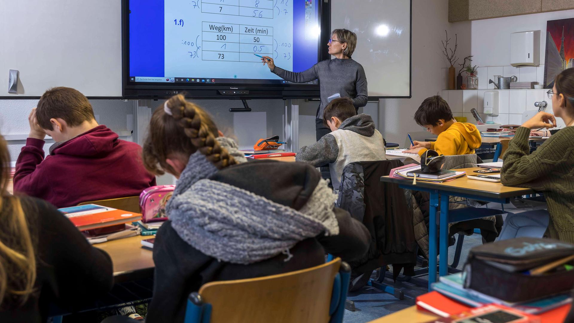 Eine Lehrerin steht im Klassenzimmer an einem interaktiven Whiteboard und erklärt eine Mathematik-Aufgabe. Mehrere Schülerinnen und Schüler sitzen an ihren Plätzen und schreiben mit. Eine Lehrerin steht im Klassenzimmer an einem interaktiven Whiteboard und erklärt eine Mathematik-Aufgabe. Mehrere Schülerinnen und Schüler sitzen an ihren Plätzen und schreiben mit.