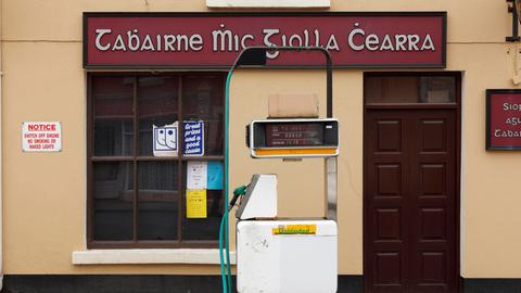 Das Schild einer Tankstelle und eines Pubs sind in gälischer Sprache verfasst. Im Vordergrund ist eine Zapfsäule.