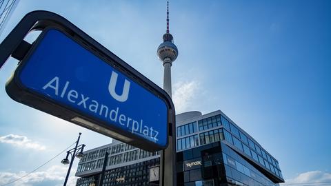 Aufnahme des U-Bahn Schildes am Bahnhof Alexanderplatz. Im Hintergrund ist vor blauem Himmel auch der Fernsehturm zu sehen.