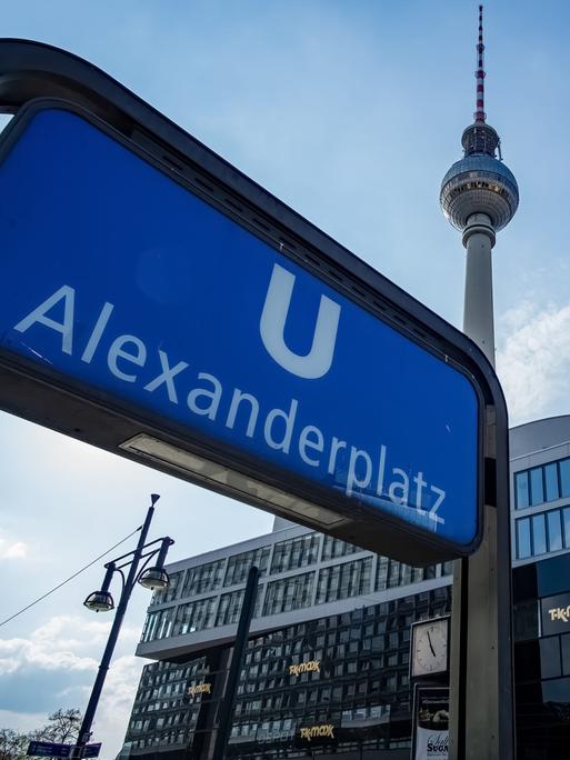 Aufnahme des U-Bahn Schildes am Bahnhof Alexanderplatz. Im Hintergrund ist vor blauem Himmel auch der Fernsehturm zu sehen.