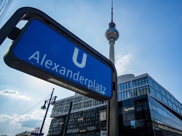 Aufnahme des U-Bahn Schildes am Bahnhof Alexanderplatz. Im Hintergrund ist vor blauem Himmel auch der Fernsehturm zu sehen.