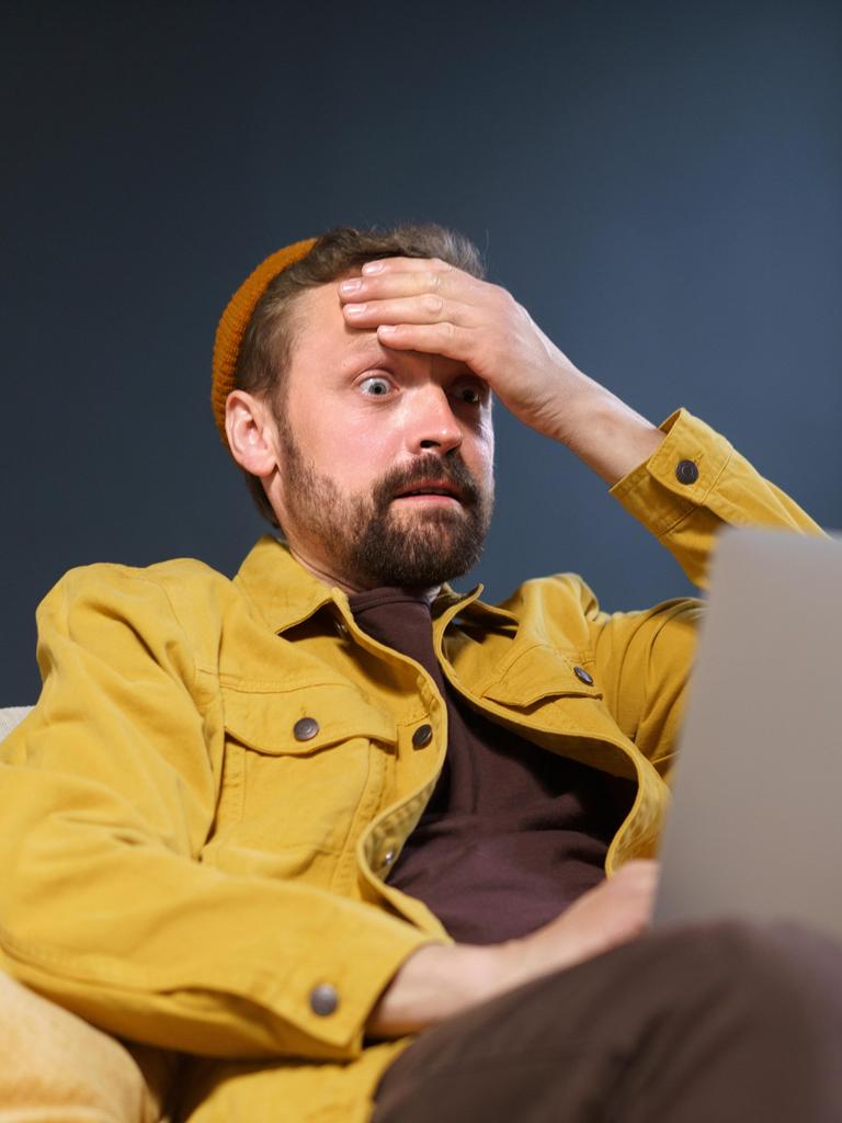 Ein Mann sitzt mit seinem Laptop auf dem Sofa und fasst sich an den Kopf. 