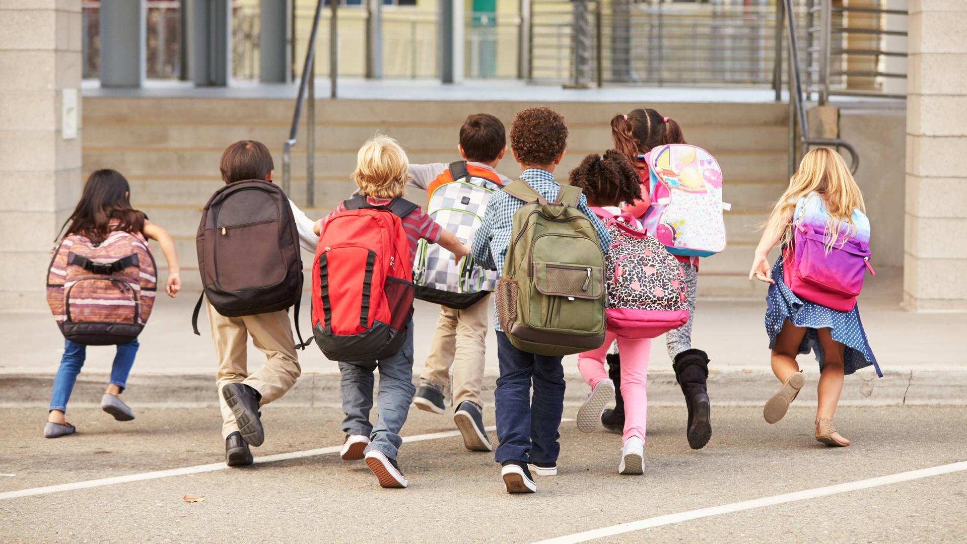 Symbolbild: Kinder mit bunten Rucksäcken laufen auf einem Schulhof in Richtung Schultor.