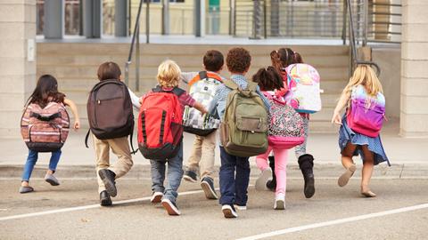 Symbolbild: Kinder mit bunten Rucksäcken laufen auf einem Schulhof in Richtung Schultor.