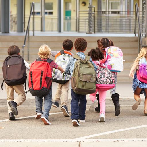 Symbolbild: Kinder mit bunten Rucksäcken laufen auf einem Schulhof in Richtung Schultor.