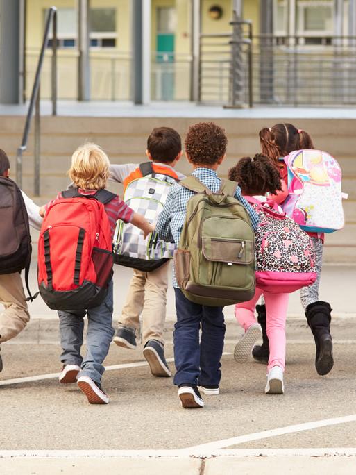 Symbolbild: Kinder mit bunten Rucksäcken laufen auf einem Schulhof in Richtung Schultor.