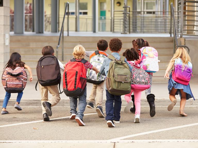 Symbolbild: Kinder mit bunten Rucksäcken laufen auf einem Schulhof in Richtung Schultor.