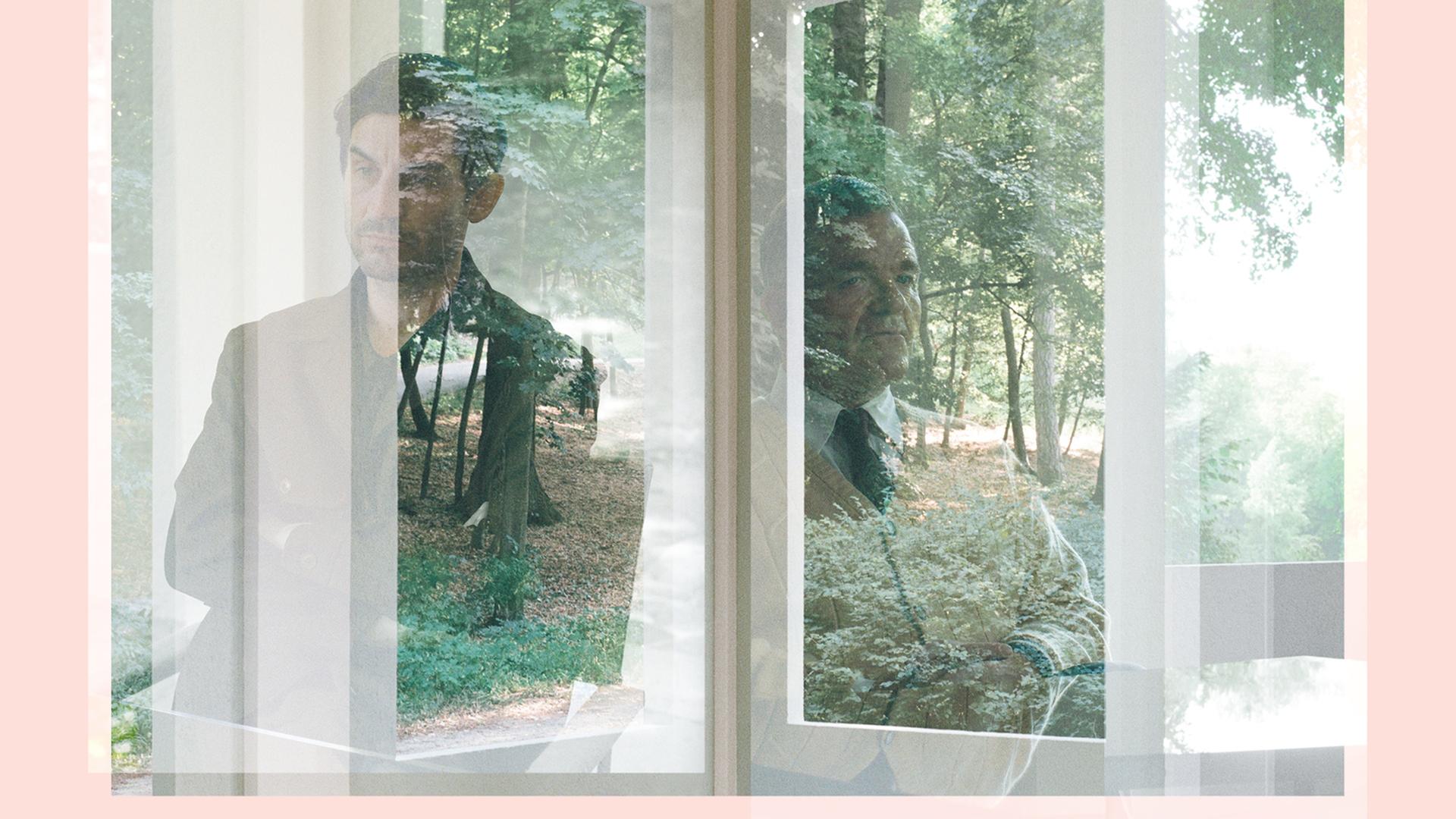Filmstill aus "De Facto" von Selma Doborac. Ein jüngerer und ein älterer Mann spiegel sich in den Scheiben eines Eckfensters.