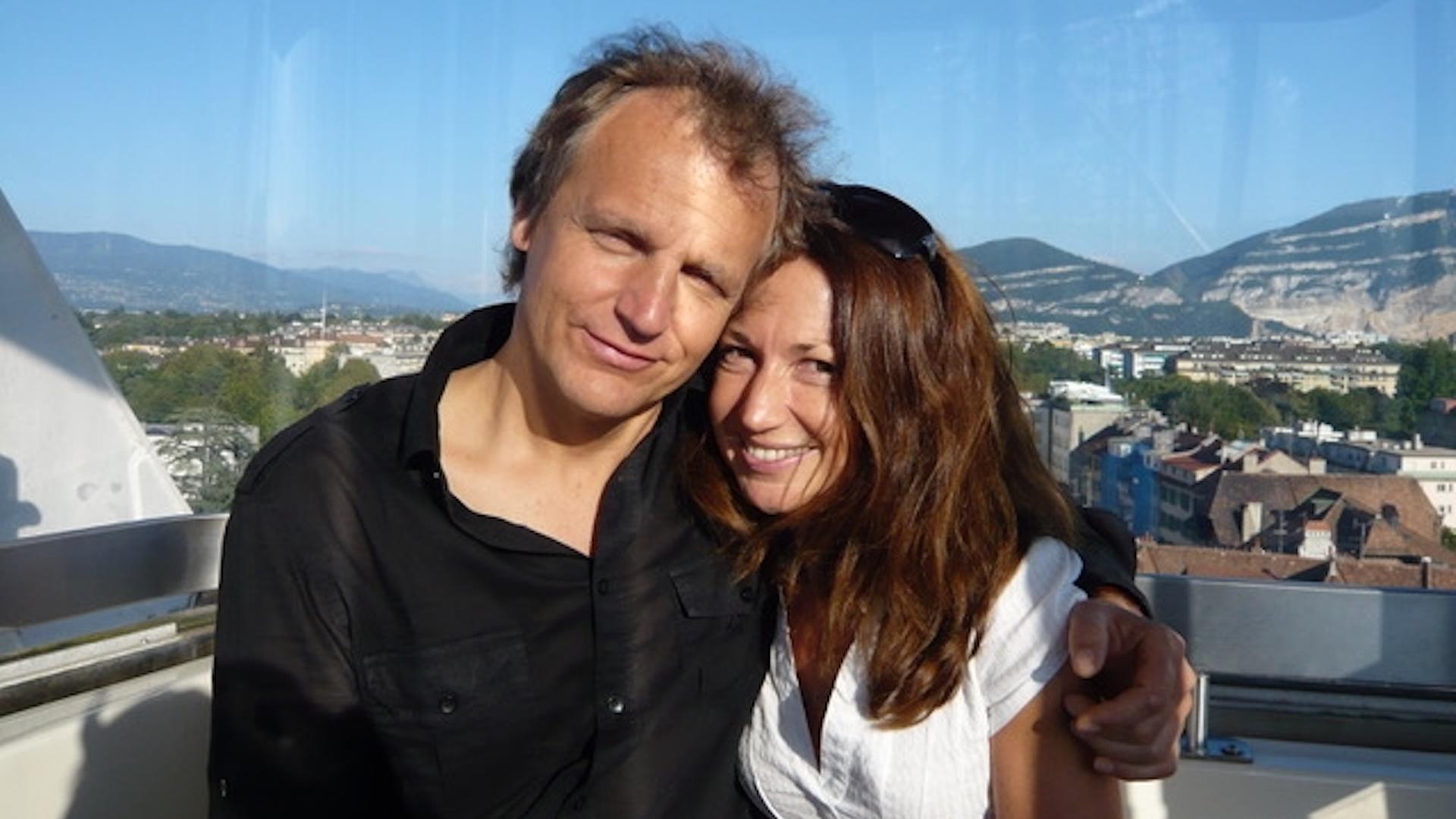 Ein Mann und eine Frau halten einander auf einem Balkon stehend in den Armen und lächeln in die Kamera.