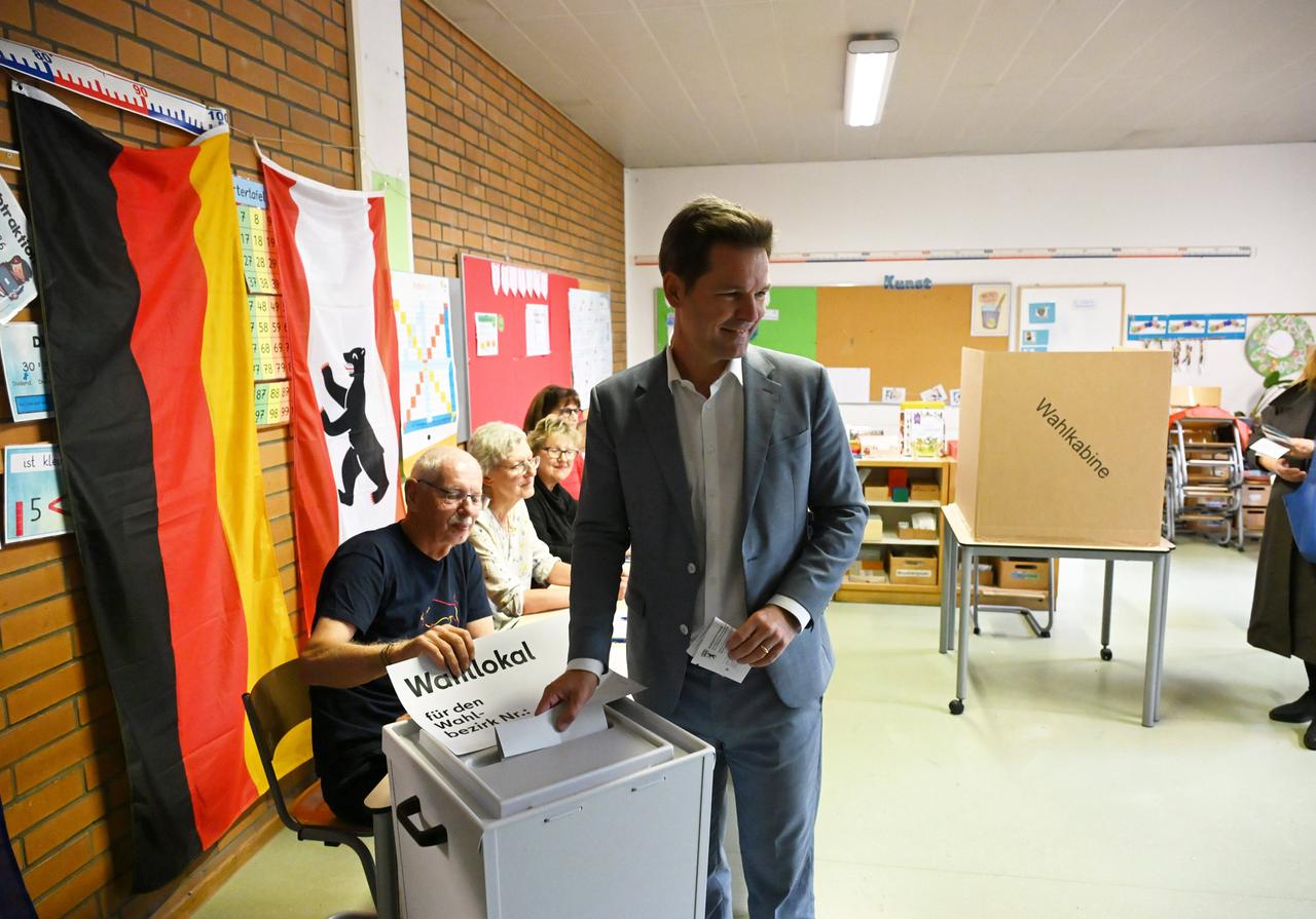 Ein Mann im grauen Anzug steckt einen Stimmzettel in die Wahlurne. Im Hintergrund ist eine Wahlkabine zu sehen. An der Wand hängt eine Deutschland- sowie eine Berliner Flagge.