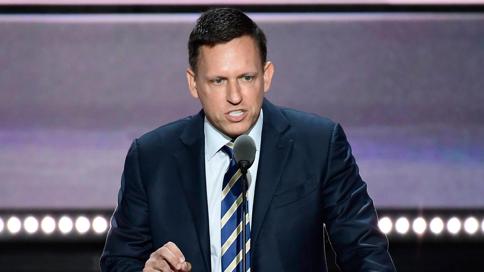 Der PayPal-Mitbegründer Peter Thiel vor einem Mikrofon. Er schaut ernst und gestikuliert mit seiner rechten Hand. Er trägt einen dunklen Anzug und Krawatte.