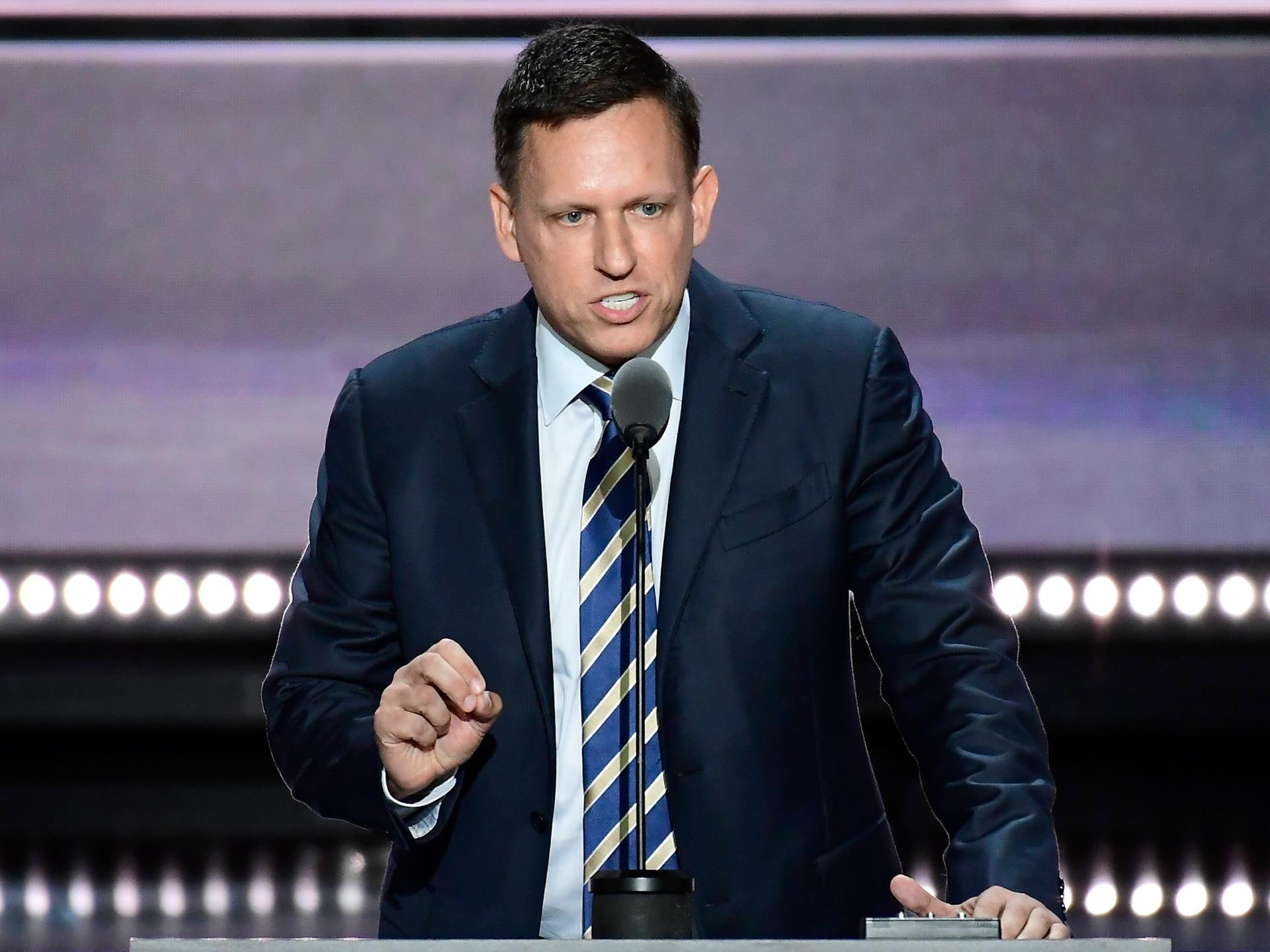 Der PayPal-Mitbegründer Peter Thiel vor einem Mikrofon. Er schaut ernst und gestikuliert mit seiner rechten Hand. Er trägt einen dunklen Anzug und Krawatte.