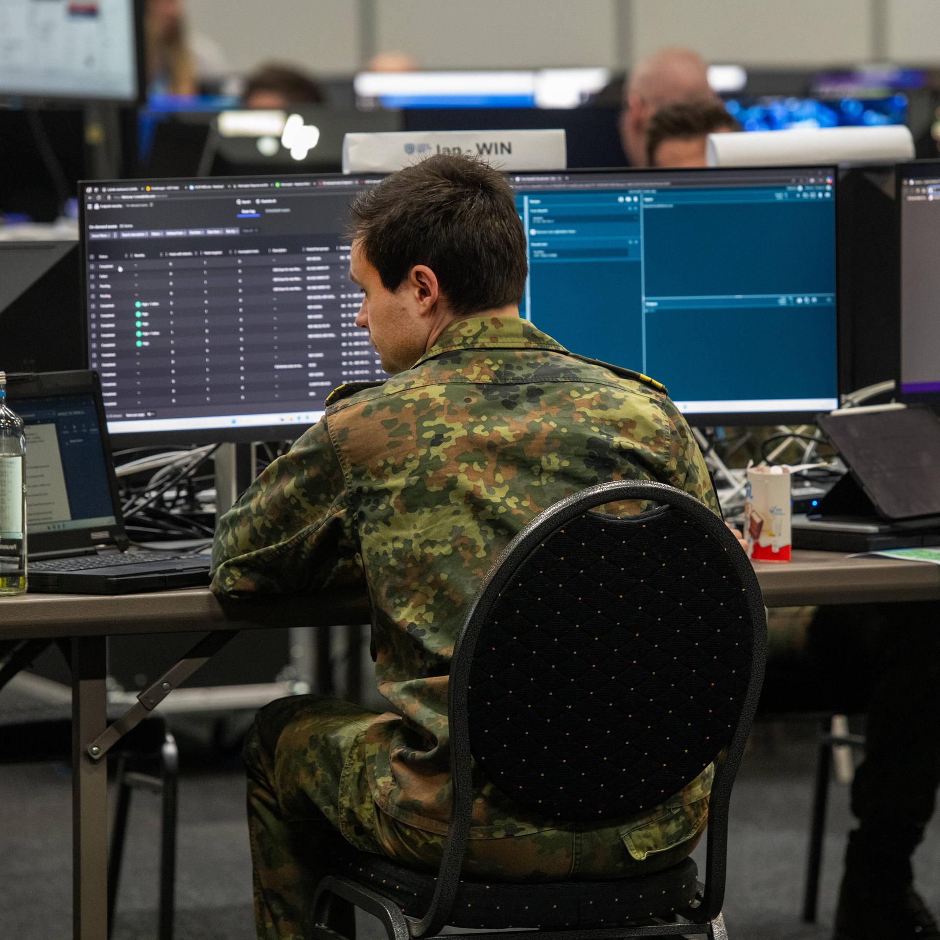 Ein Mann in Bundeswehr-Uniform sitzt vor mehreren Bildschirmen. Im Hintergrund arbeiten weitere Soldaten an Computern.