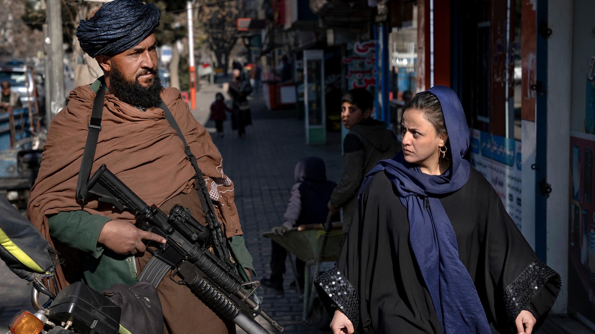 Ein Taliban-Kämpfer steht Wache, während eine Frau vorbeiläuft. Der Taliban hat eine Waffe in der Hand. Die Fraue trägt locker ein Tuch über den Haaren und schaut den Taliban böse an. 