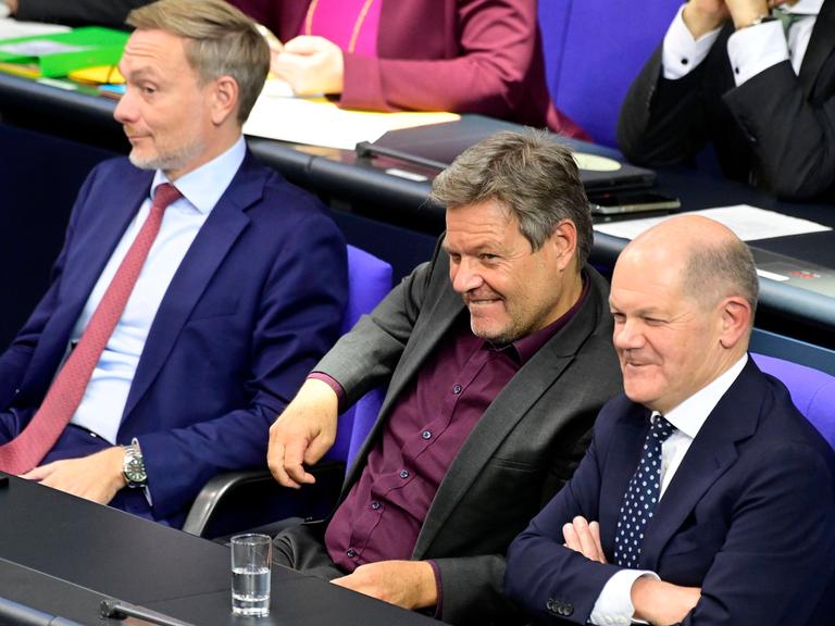 Christian Lindner, Robert Habeck und Olaf Scholz sitzen im Bundestag nebeneinander und grinsen