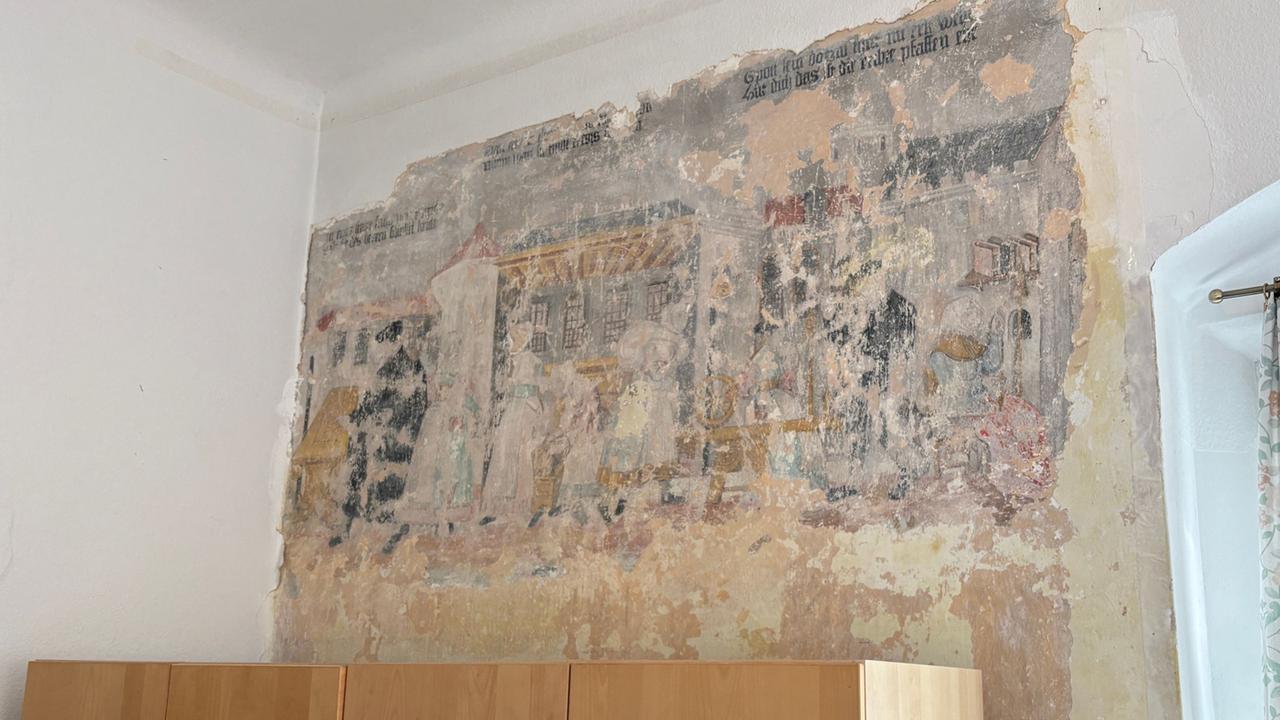 Das Bild zeigt fragmentarisch eine Kalk-Secco-Wandmalerei in restaurierungsbedürftigem Zustand, die vermutlich Szenen der frühneuzeitlichen Textilverarbeitung darstellt.