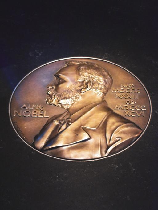Auf einem schwarzem Untergrund glänzt eine goldene Münze, die das Konterfei von Alfred Nobel zeigt.