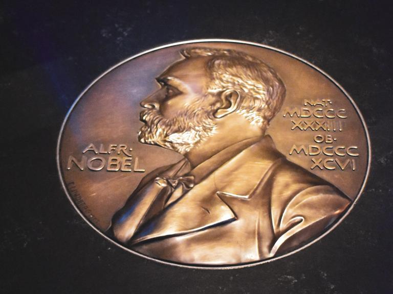 Auf einem schwarzem Untergrund glänzt eine goldene Münze, die das Konterfei von Alfred Nobel zeigt.