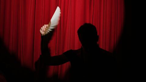 Eine Theaterfigur hält einen Fächer in einen Lichtkegel vor einem roten, geschlossenen Bühnenvorhang aus Samt. 