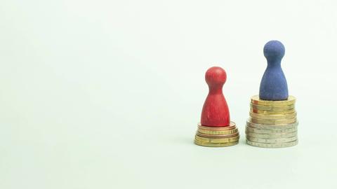 Eine rote Spielfigur steht auf einem kleineren Münzstapel, eine blaue Spielfigur auf einem größeren, um die unterschiedlichen Einkommensniveaus von Männern und Frauen zu visualisieren.