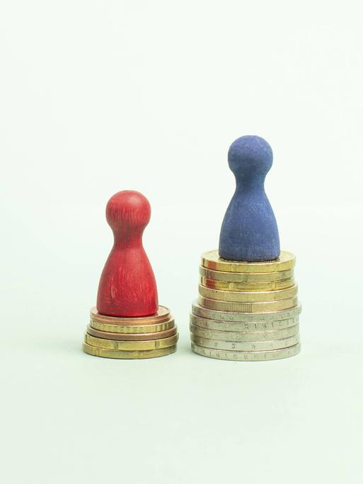 Eine rote Spielfigur steht auf einem kleineren Münzstapel, eine blaue Spielfigur auf einem größeren, um die unterschiedlichen Einkommensniveaus von Männern und Frauen zu visualisieren.