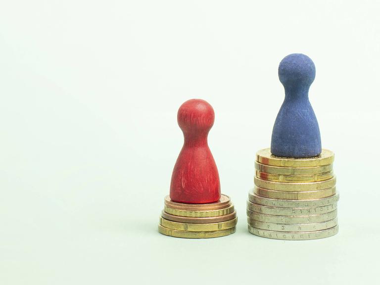 Eine rote Spielfigur steht auf einem kleineren Münzstapel, eine blaue Spielfigur auf einem größeren, um die unterschiedlichen Einkommensniveaus von Männern und Frauen zu visualisieren.