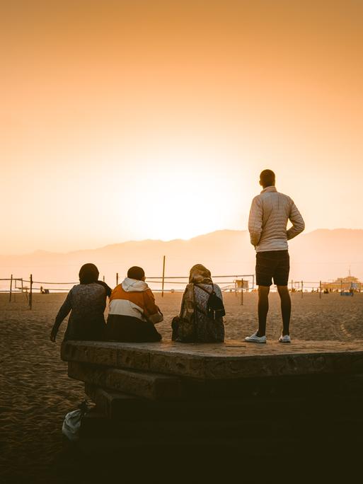 Eine vierköpfige Gruppe steht mit dem Rücken zur Kamera an einem weitläufig und sonst menschenleeren Strand und betrachtet den Sonnenuntergang.