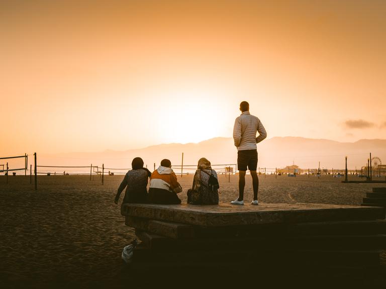 Eine vierköpfige Gruppe steht mit dem Rücken zur Kamera an einem weitläufig und sonst menschenleeren Strand und betrachtet den Sonnenuntergang.