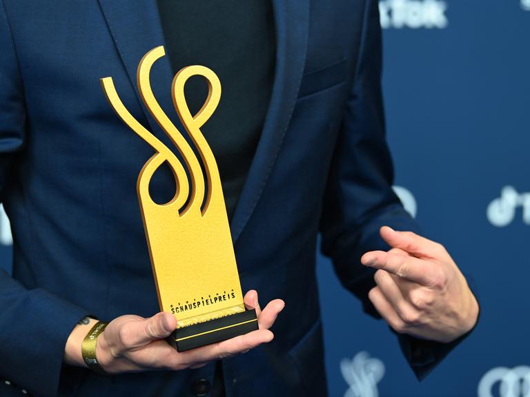Eine Hand hält den Deutschen Schauspielpreis, die andere zeigt darauf.