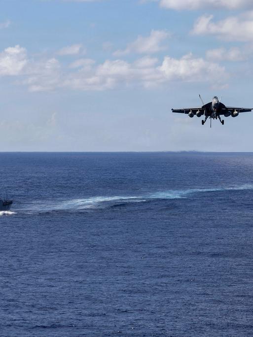 Zwei militärische Schiffe fahren auf dem offenen Meer, dazwischen setzt ein Kampfflugzeug zum Landeanflug an.