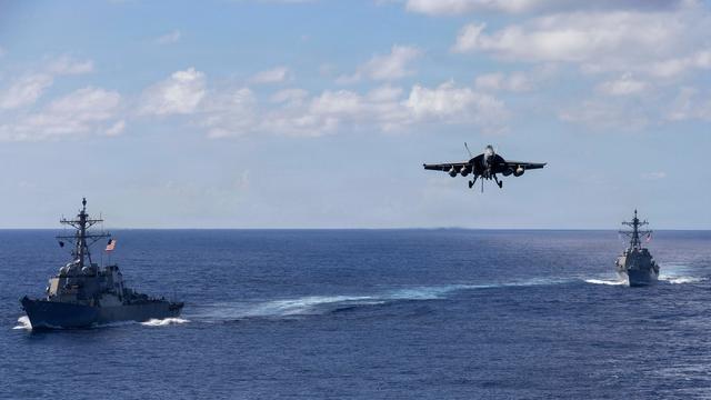 Zwei militärische Schiffe fahren auf dem offenen Meer, dazwischen setzt ein Kampfflugzeug zum Landeanflug an.