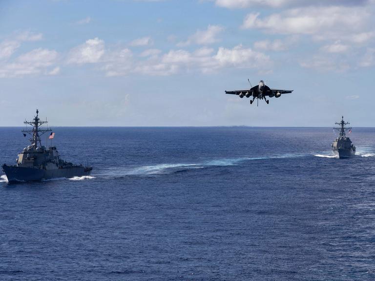Zwei militärische Schiffe fahren auf dem offenen Meer, dazwischen setzt ein Kampfflugzeug zum Landeanflug an.